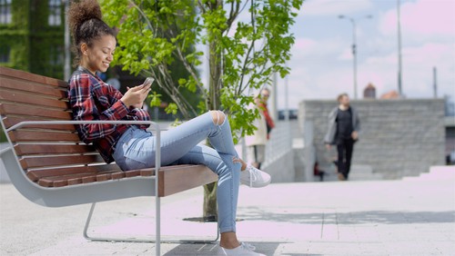 Pretty teenage girl using her mobile phone while sitting on wooden bench. Casual style - jeans and checkered shirt.